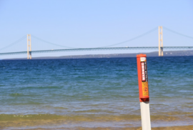 A warning marker sits at the water's edge with the Mackinac Bridge in the background