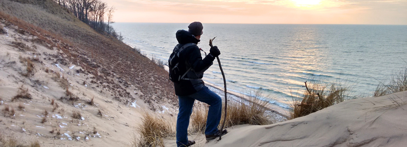 A man stands atop Michigan's sand dunes overlooking one of our Great Lakes, holding a walking stick