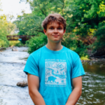 Tommy Skinner, 2023 Petoskey Prize recipient, stands smiling in front of a flowing river in Petoskey.