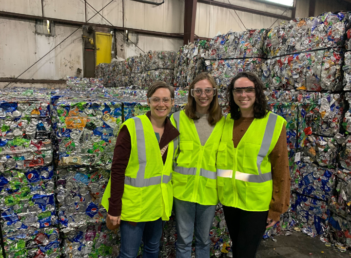 Three women wearing safety goggles and vests stand smiling in front of organized bales of recycled bottles