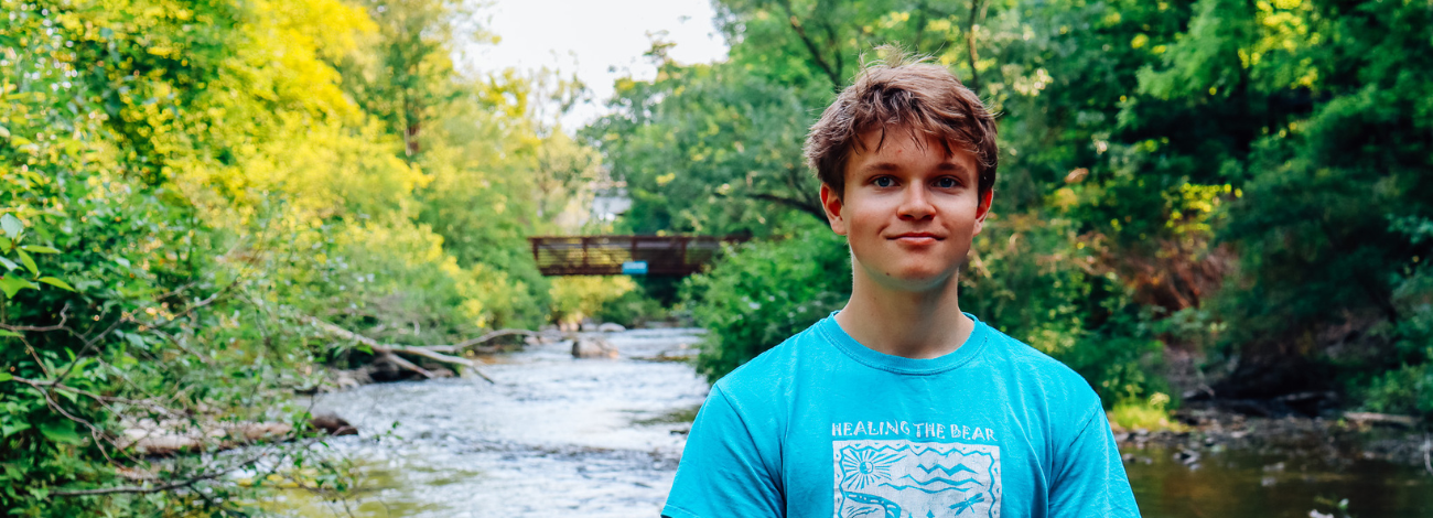 Tommy Skinner, 2023 Petoskey Prize recipient, stands smiling in front of a flowing river in Petoskey.