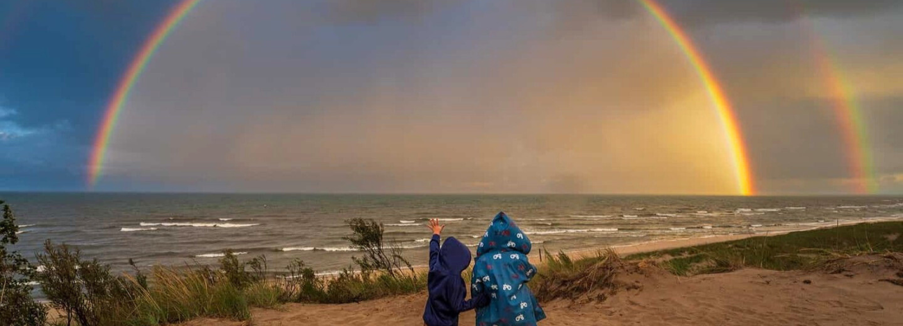 Two young boys stand in raincoats on a Michigan beach, one points up at the sky where a double rainbow has appeared over the water.