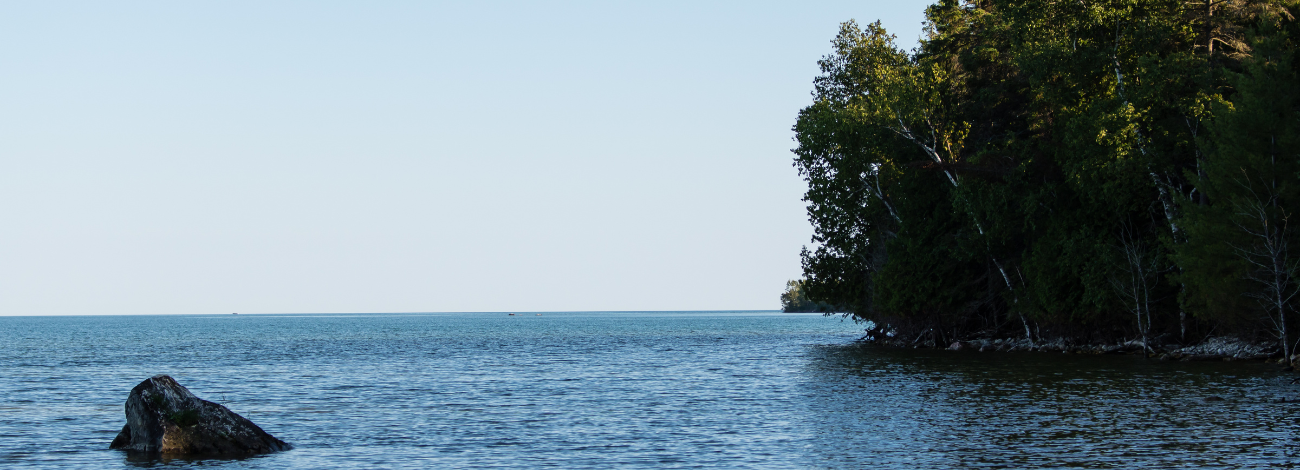 A large rock sits in Lake Huron's peaceful waters; trees cover the shoreline to the right.