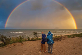 Two young boys stand in raincoats on a Michigan beach, one points up at the sky where a double rainbow has appeared over the water.