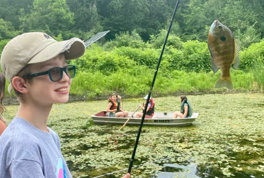 A boy shows off a fish he caught with his fishing rod while a group of girls paddle behind him on a lake. (Photo courtesy of Izaac Walton League - Dwight Lydell Chapter)
