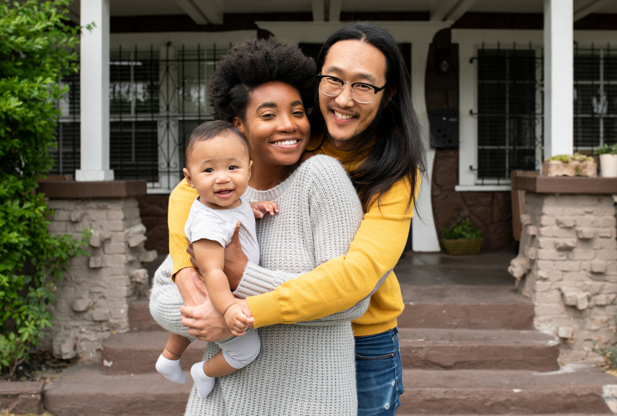 A young family hugs each other for a photo in front of their home's porch steps (Photo via Adobe Stock)
