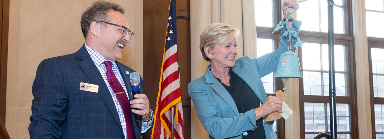 Former Michigan Governor Jennifer Granholm (right) rings the blue Milliken Bell for Distinguished Leadership. MI Environmental Council president & CEO (left) smiles and looks on as he holds the microphone.